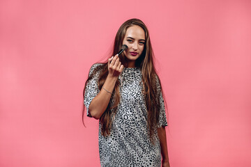 Cute young woman with long hair in casual outfit holding a makeup brush on her velvet skin against a pink background. Make up concept.