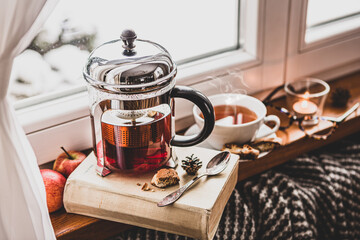 Winter relax at cozy home. Glass teapot with hot tea and book by the window.