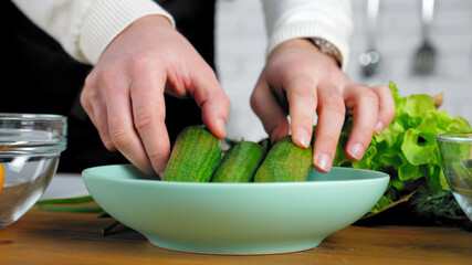 Professional chef man in black apron standing near table prepares ingredients vegetables for salad at home kitchen. Close up hands put fresh food cucumbers in plate