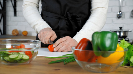 Professional chef cook man in black apron standing near table sliced fresh cherry tomato on cutting board at home kitchen. Middle shot male hands food vegetables ingredients for salad dish