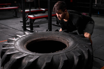 spotsman pushing drawn tire to build strength at modern fitness center. Male in sportswear is...