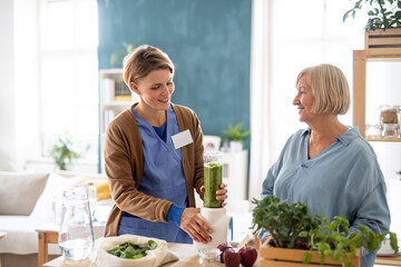 Senior woman with caregiver or healthcare worker indoors, preparing healthy smoothie.