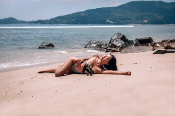Fototapeta na wymiar Young sexual woman with long legs and a charming body is lying on the golden sand against the background of beating waves on the rocks