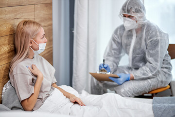 doctor in protective suit asks female patient about state of health, professional doctor taking notes, coronavirus covid-19. stay at home concept during Coronavirus. focus on sick female