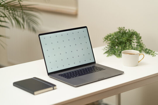 Workspace with laptop, green plants in the background and black notebook, white coffee in golden cup on the white table.