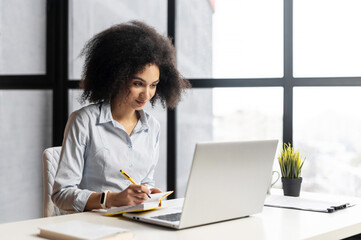 Positive African American woman studying or working in the office, sitting at the desk, using the laptop for distance video communication, watching webinar, having a meeting online, and making notes