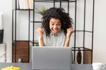Happy excited African American girl raised fists celebrating success, making yes gesture, looking at the laptop screen, passed exam test with a high score, got promoted, won a lottery, victory concept