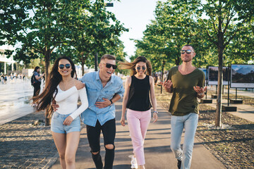 Happy multiethnic friends in casual wear and sunglasses smiling and looking at each other while walking in sunny park together