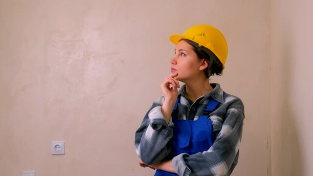 A young construction worker in a yellow hard hat and blue uniform stands against the stucco wall and looks around thoughtfully
