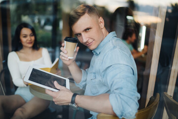 Portrait of handsome Caucasian blogger holding takeaway caffeine beverage and modern digital tablet in hands and looking at camera during pastime with blurred girlfriend, window view on millennial man