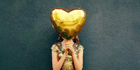 Woman covering her head with big golden heart shaped balloon on a dark background