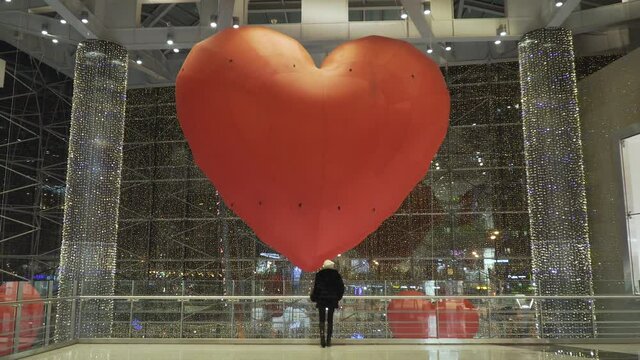 portrait of a young caucasian girl in a black fur coat and a pink hat in a shopping mall looks at a huge heart-shaped balloon on valentine's day