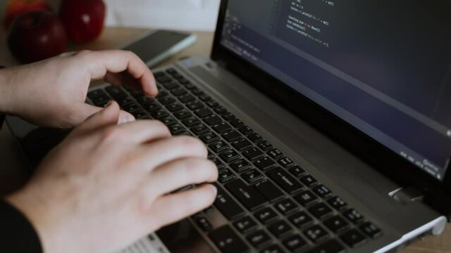 Men's hands press the keys of a laptop keyboard. A man writes a program. The developer is coding. The program code is visible on the screen. Work at home
