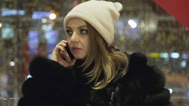 portrait of a young caucasian girl in a black fur coat and a pink hat in a shopping mall against the background of a huge heart-shaped balloon on valentine's day. she is talking on the phone