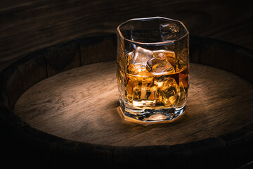 Glass of drink with a strong drink with ice on the background of an oak barrel with alcohol. Background with whiskey with ice in a glass close-up with copy space.