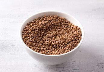 Raw buckwheat in a white ceramic bowl on a light gray textured background, horizontal, close up