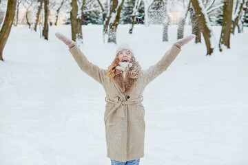 A happy young blonde woman throws up snow and enjoys the snowfall. Coat with fur. Winter park with snow.