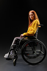 Fototapeta na wymiar sad disabled woman in a wheelchair, looking at camera depressed. redhead female in yellow casual shirt sits isolated on black background. health and people concept