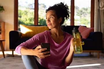 Caucasian woman holding water bottle and smartphone taking break from exercising at home