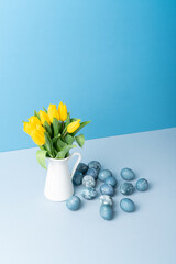 Bouquet of yellow fresh tulips in white jug, painted Easter eggs on blue background. Place for text.