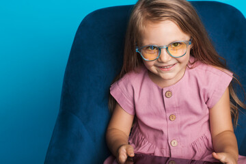 Close up photo of a young girl smiling at camera in yellow glasses while sitting in armchair on a blue studio wall near free space