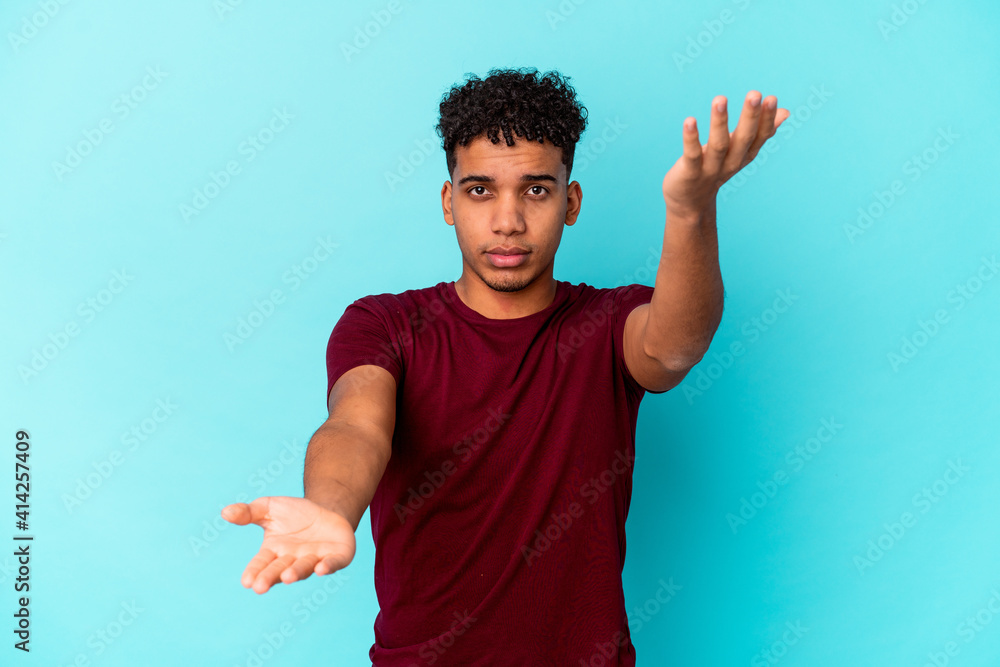 Wall mural young african american curly man isolated on blue makes scale with arms, feels happy and confident.