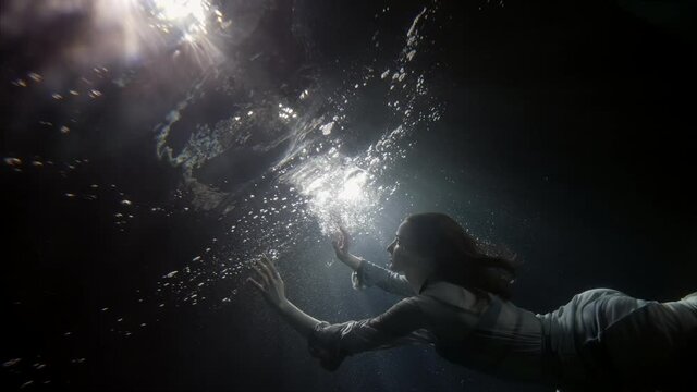 A beautiful woman in a flowing dress floats under the water on a dark background, she swims to the surface of the water and comes up, creating a lot of bubbles.