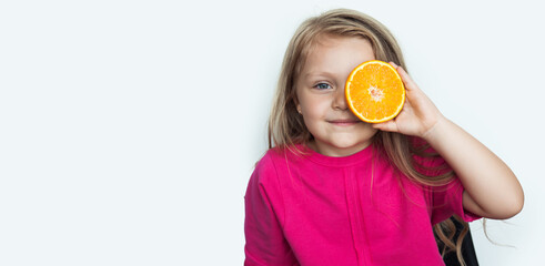 Adorable small girl is covering her eye with an orange smiling at camera on a white studio wall with free space