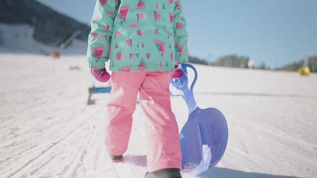 Slow motion close up - Little girl walking uphill in snow park to go sledding. Fun family time