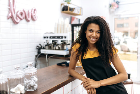 Photo of a friendly waitress wearing uniform, african american female barista, with curly hair in black apron standing near the bar counter, looks and smiles at the camera