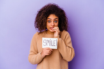 Young African American woman holding a Smile placard isolated on purple background biting fingernails, nervous and very anxious.