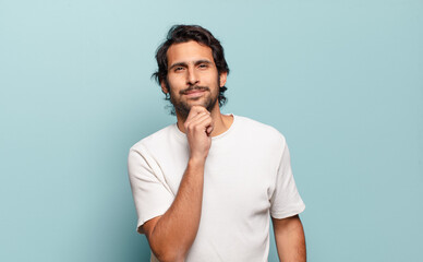 young handsome indian man looking happy and smiling with hand on chin, wondering or asking a...
