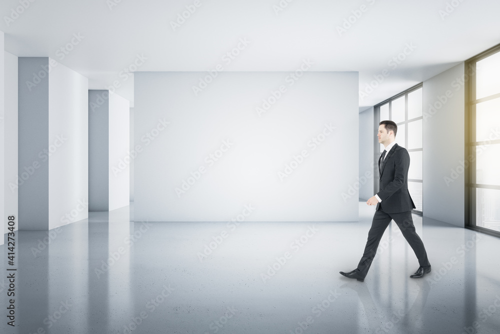 Poster young businessman walking in light spacious minimalistic room with blank light walls and windows to 