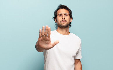 young handsome indian man looking serious, stern, displeased and angry showing open palm making...