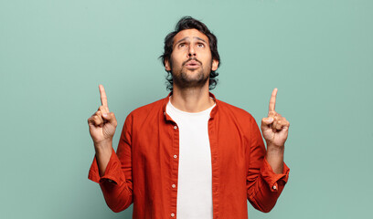 young handsome indian man feeling awed and open mouthed pointing upwards with a shocked and...