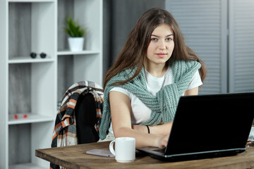 A long-haired brunette is preparing for a test exam doing homework at home sitting at a table at a laptop posing for the camera. The concept of self-education, distance learning.