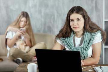 A long-haired brunette is preparing for a test exam at home sitting at a table with a laptop looking into the camera, in the background her friend with a phone in her hands