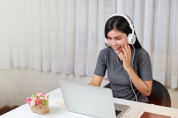 Young smart and active asian female student sitting at table with headset, using computer and taking note for studying online with happy smiling face (new normal concept)