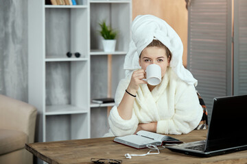 A young girl in a bathrobe and with a towel on her head sits with a cup in her hands at a table with a laptop and looks at the camera in the background of a bookcase. Student everyday life.