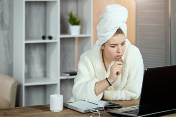 An emotional, relaxed young beautiful girl in a bathrobe and a towel on her head, sitting at a table watching movies or TV shows on a laptop and eating chocolate. Chill out and leisure concept.