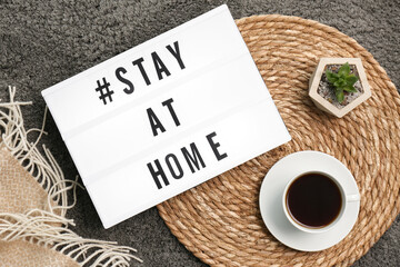 Cup of coffee, houseplant and lightbox with hashtag STAY AT HOME on grey carpet, flat lay. Message to promote self-isolation during COVID‑19 pandemic