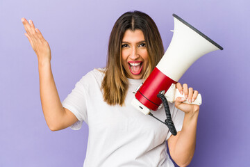 Young indian woman holding a megaphone isolated receiving a pleasant surprise, excited and raising hands.