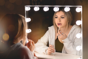 Young teenage girl applying lip gloss in front of vanity mirror with lights