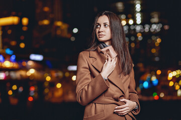 Close up portrait of a lovely pleasant dark-haired lady in a coat posing against the backdrop of the bright lights of the night city looking into the distance from the belts. Night city concept