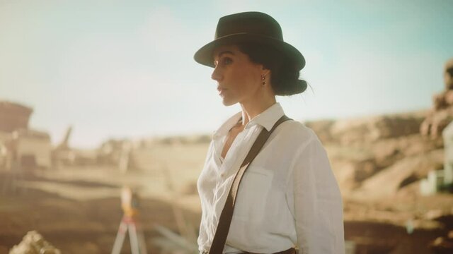 Portrait of Beautiful Female Adventurer Wearing a Hat Posing and Looking at Camera. Stylish Great Archaeologist Standing with Ancient Civilization, Fossil Remains Archeological Site in Background