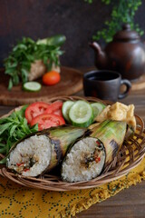 nasi bakar or baked rice with dishes wrapped in leaves served with vegetables in a woven plate on the table