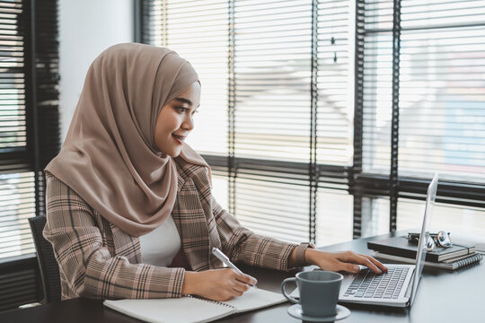 Beautiful Asian Muslim Business Woman Brown Hijab Sitting And Working With Laptop Computer At Modern Office.