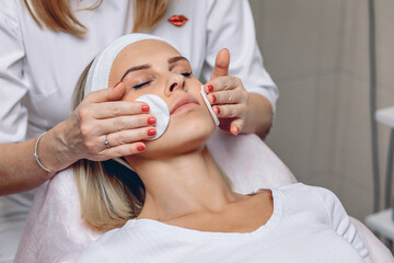 Master beautician wiping the face of a young and beautiful client with cotton pads. Cosmetic...