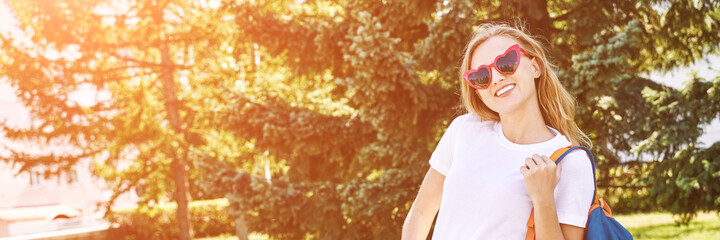 Young woman with bright backpack. Local tourist. Summer sunny day. Green background. Staycation weekend. Female person in love eyeglasses and white tshirt. Smiling outdoors portrait. Horizontal banner