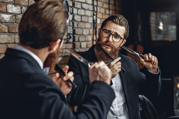 Attractive charming male barber in glasses, with a mustache and a beard, looking in the mirror and holding a comb and scissors next to his face. Barbershop concept
